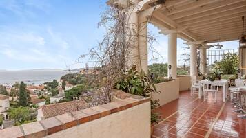 A covered outdoor area with a terrace, potted plants, and a view of the sea and buildings.