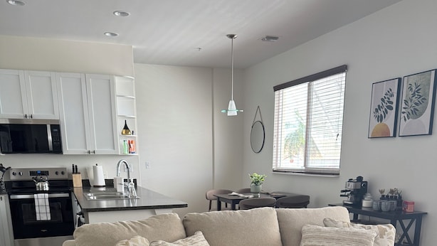 Un salon moderne avec un canapé d’angle beige, une table basse ronde et un coin cuisine doté de meubles blancs et d’une cuisinière noire.