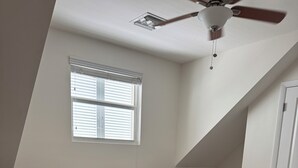 Une chambre à coucher avec un lit, une table de chevet, une lampe, une fenêtre et un ventilateur de plafond.
