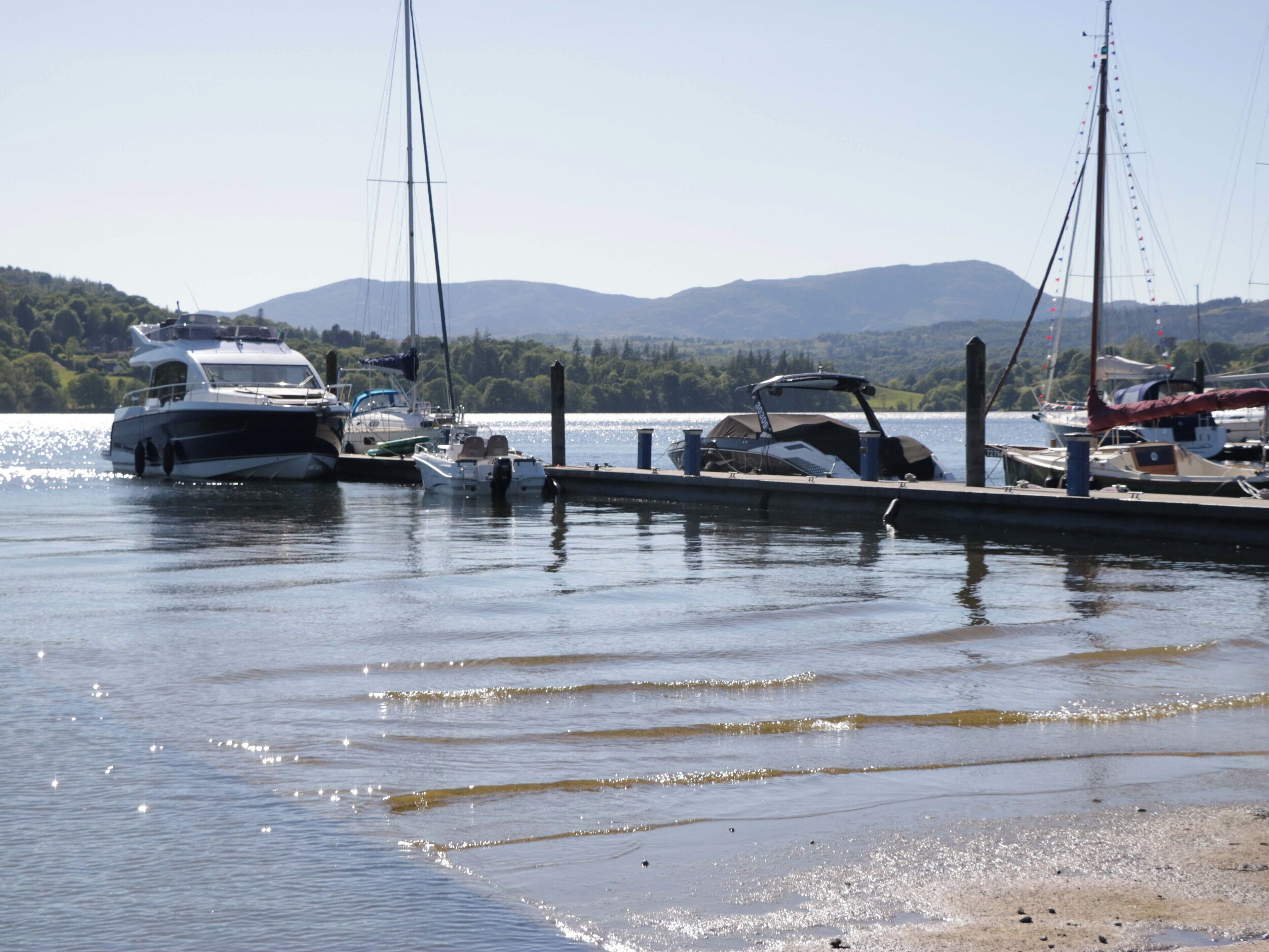 A marina with boats docked, calm water, and a hilly landscape in the background.