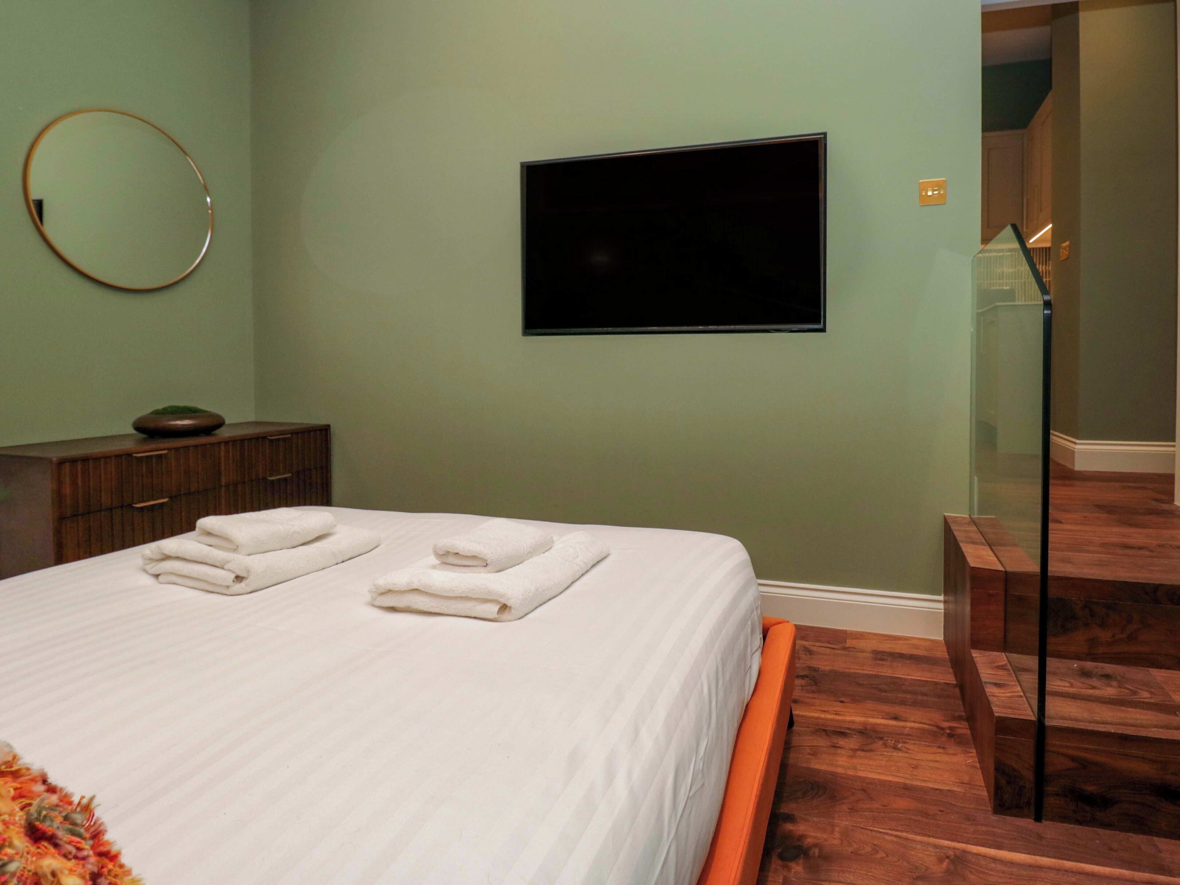 A neatly made bed with two folded white towels, a wooden dresser, a round mirror, and a flat-screen TV mounted on the wall.