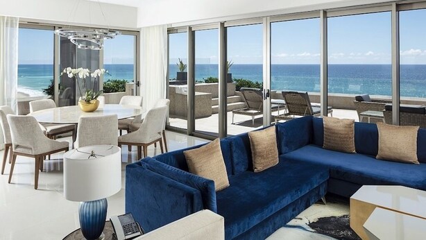A modern living room with a large blue sectional sofa, a dining area, and a view of the ocean. - 4,198-square-foot oceanfront suite is chic and modern (Aurora)