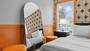 Hotelkamer met een grote spiegel, een bed met wit bedlinnen, een oranje poef, een tafeltje met een lamp en een kamer met een patroon op de muur.