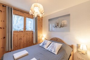 A room with a bed, wooden walls, a window with curtains, a wall-mounted lamp, and a framed picture. - Cosy Apartment in Central Samoëns with Mountain View, Balcony, Near the Slopes (Samoëns)