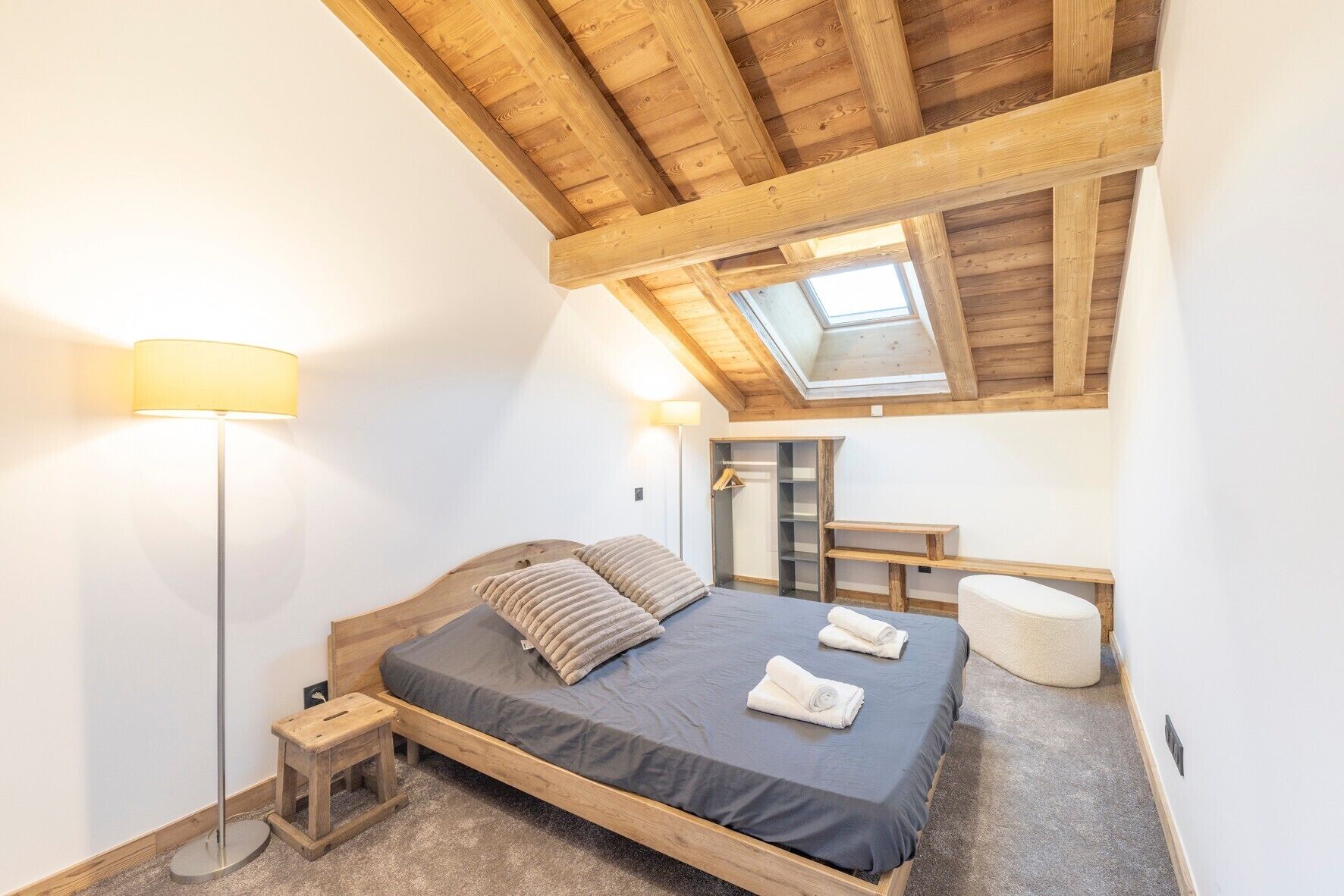 A bedroom with a sloped ceiling, a skylight, a bed with pillows, a bedside table, a lamp, and a bench.