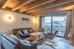 Ein gemütliches Wohnzimmer mit einem Ledersofa, einem Holztisch und einem Schaukelstuhl. Ein großes Fenster bietet einen Blick nach draußen.