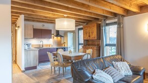 A modern kitchen with wooden cabinets, a dining area with a table and chairs, and a leather sofa with a throw pillow. - Apartment 'Morillon – New – 2 min from the slopes' with mountain view, private terrace (Morillon)