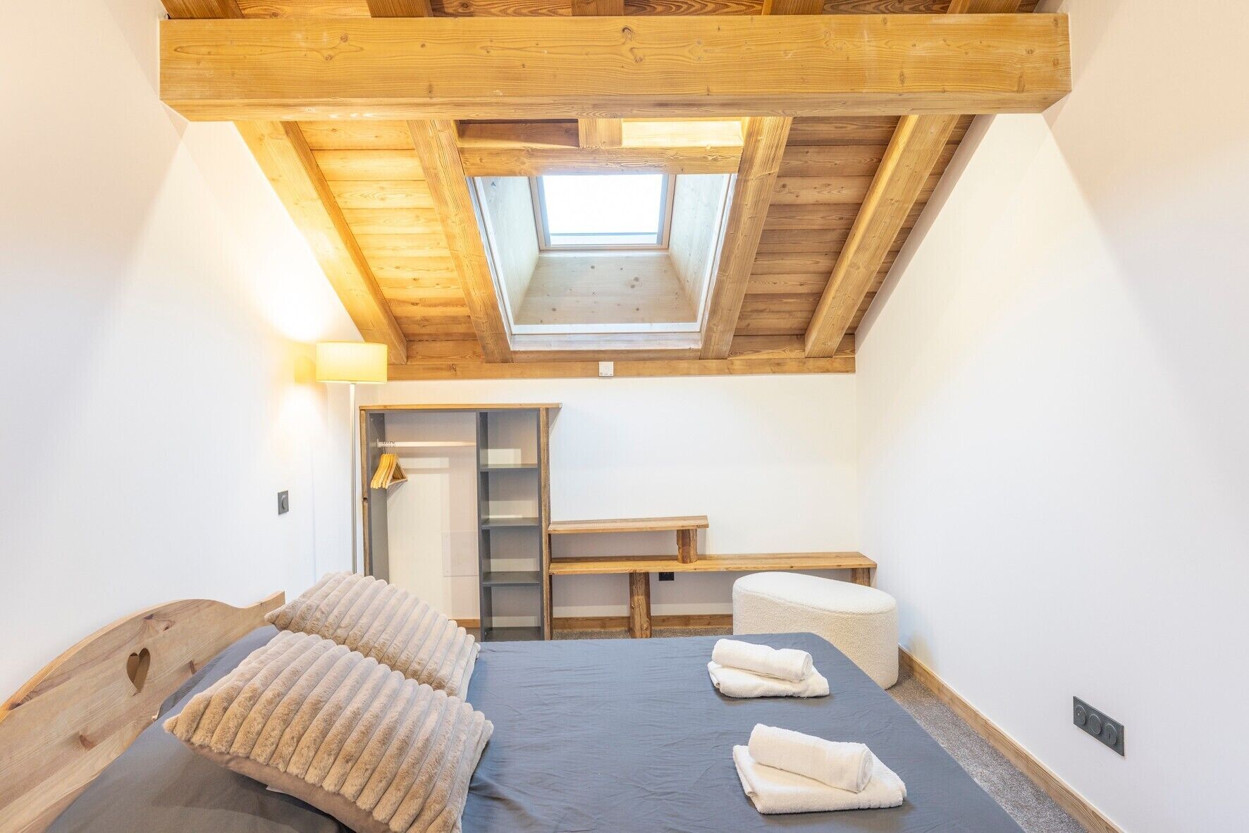 Een slaapkamer met dakraam, een bed met kussens, een nachtkastje, een bankje en een kledingkast.