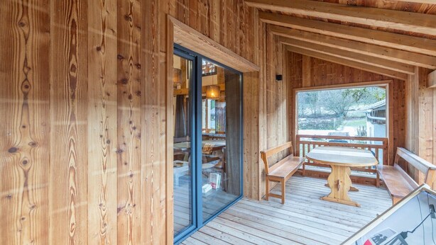 Une cabane en bois avec une porte en verre donnant sur un espace intérieur, une terrasse en bois avec une table et des bancs, et une vue sur l’extérieur.