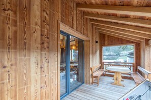 Una cabaña de madera con un ventanal de vidrio que da a un espacio interior, una terraza de madera con una mesa y bancos, y vistas al exterior.