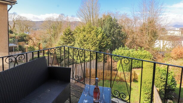 Un balcón con un sofá de mimbre y una mesa con una botella y copas, con vista a un jardín con árboles y una valla de madera.