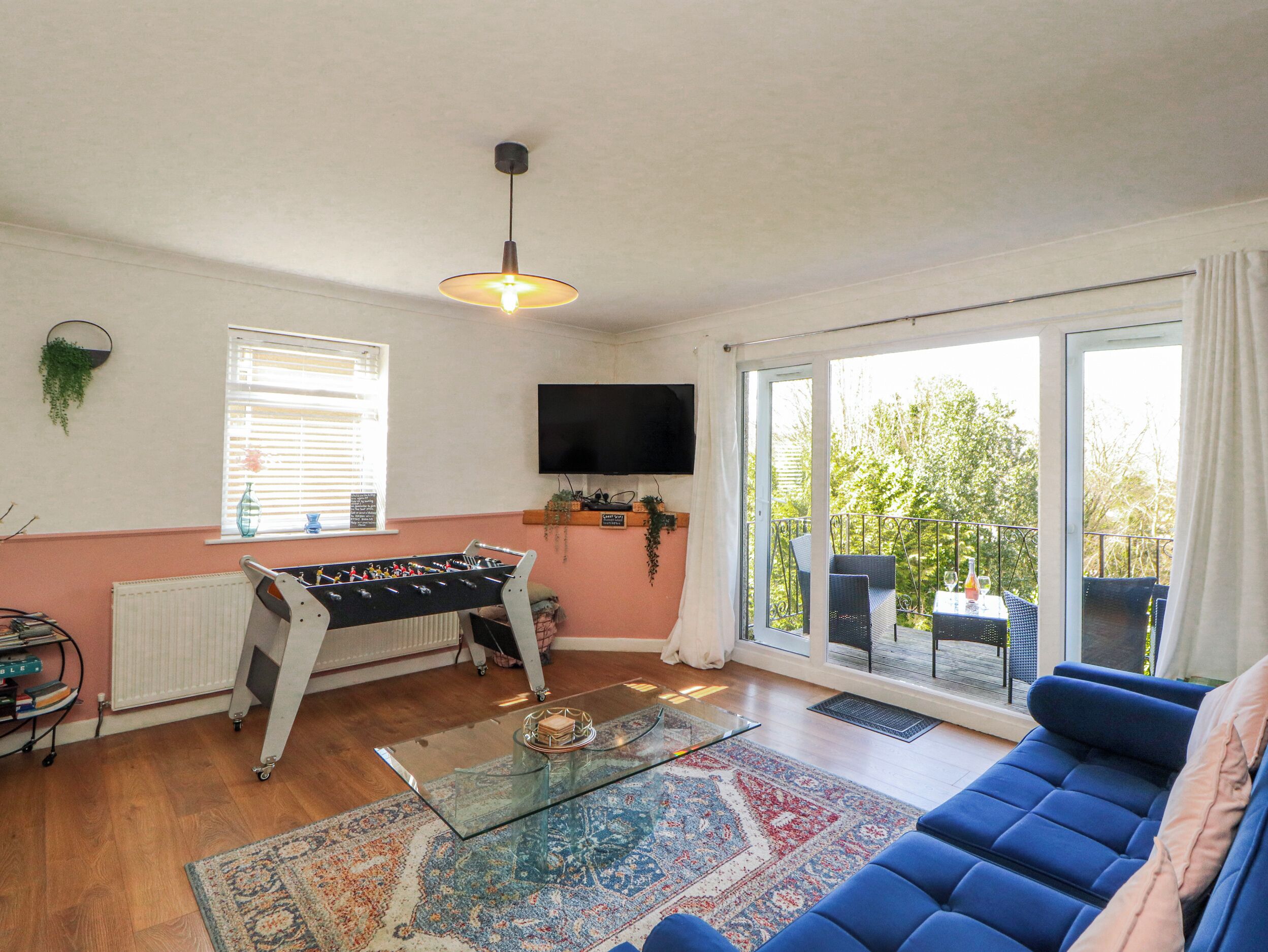 A living room with a foosball table, a glass coffee table, a sofa, a TV, and a balcony with outdoor furniture.