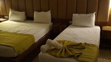 Dos camas individuales con sábanas blancas y cabeceros marrones, una mesita de noche de madera y una lámpara de pared.