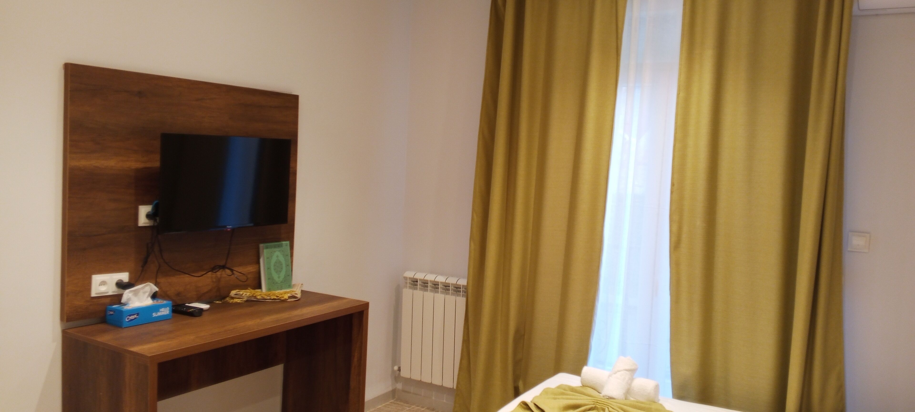 Una habitación con un escritorio de madera, un televisor montado en la pared, un radiador y una ventana con cortinas verdes.