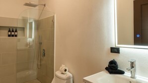 Une salle de bain moderne avec une douche à parois de verre, des toilettes blanches, une poubelle noire et un meuble-lavabo en bois avec un miroir.
