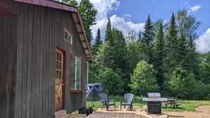 Puuinen mökki, jossa on terassi, kaksi Adirondack-tyylistä puutarhatuolia, grilli ja teltta.