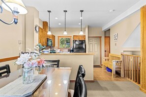 A modern kitchen with wooden cabinets, a central island, pendant lighting, and a dining area with a glass table and chairs. - Whispering Pines 3BR Townhouse Views + Hot Tub (Golden)