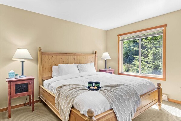 A bedroom with a wooden bed, two bedside tables with lamps, a window with a view of trees, and a tray with a mug and a book. - Whispering Pines 3BR Townhouse Views + Hot Tub (Golden)
