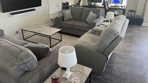 A modern living room with a large flat-screen TV, grey sofas, a coffee table, and a desk with a computer. - Perfect 3-bedroom house with AC in Fort Myers Beach (Fort Myers Beach)