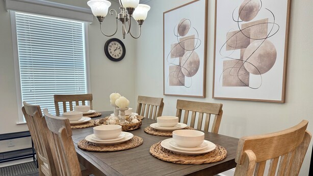 A dining room with a wooden table set for six, surrounded by wooden chairs. There are abstract paintings on the wall, a clock, and a chandelier. - Perfect 3-bedroom house with AC in Fort Myers Beach (Fort Myers Beach)