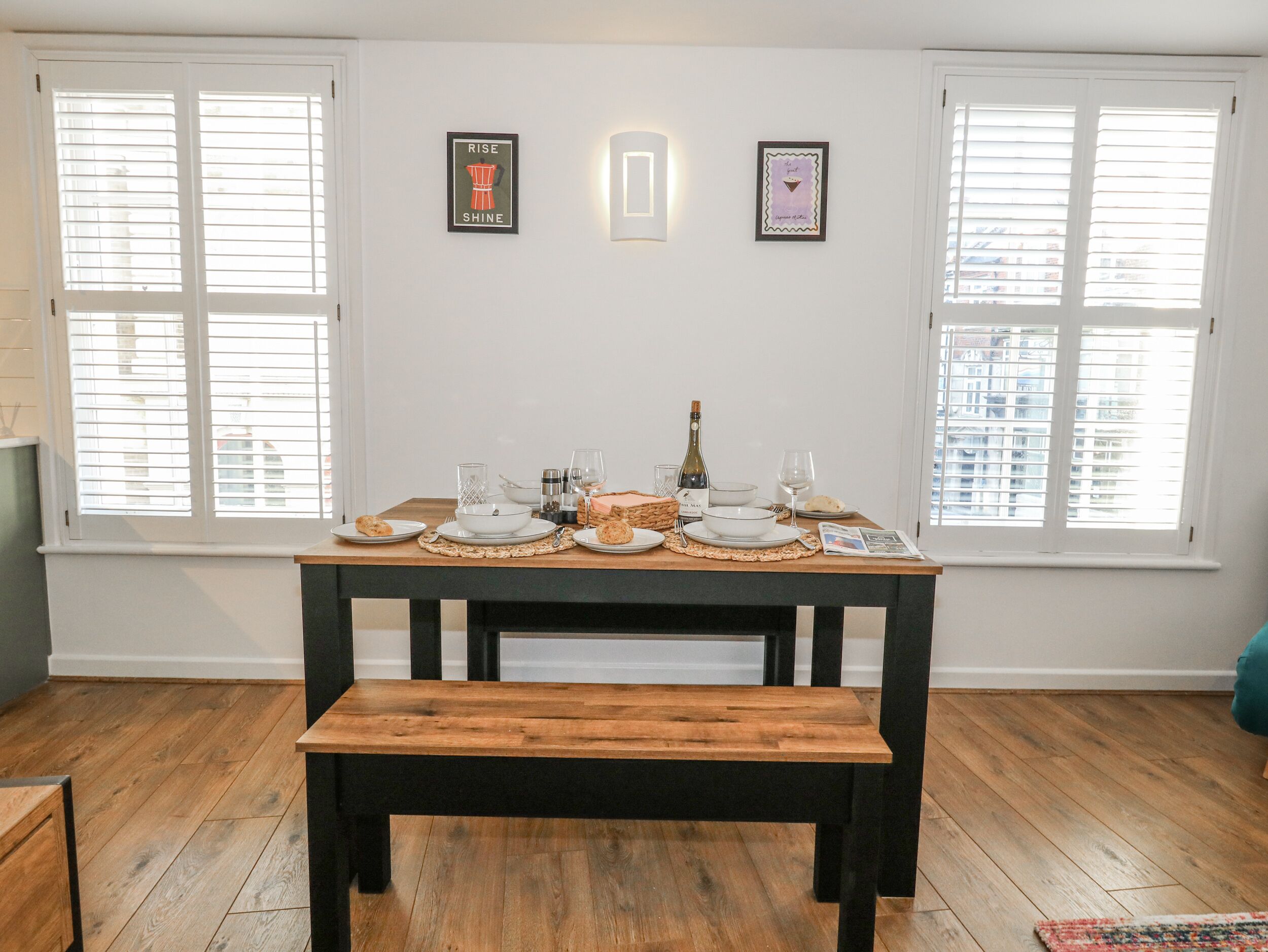 Un comedor con mesa de madera dispuesta para dos personas, flanqueado por ventanas con persianas.