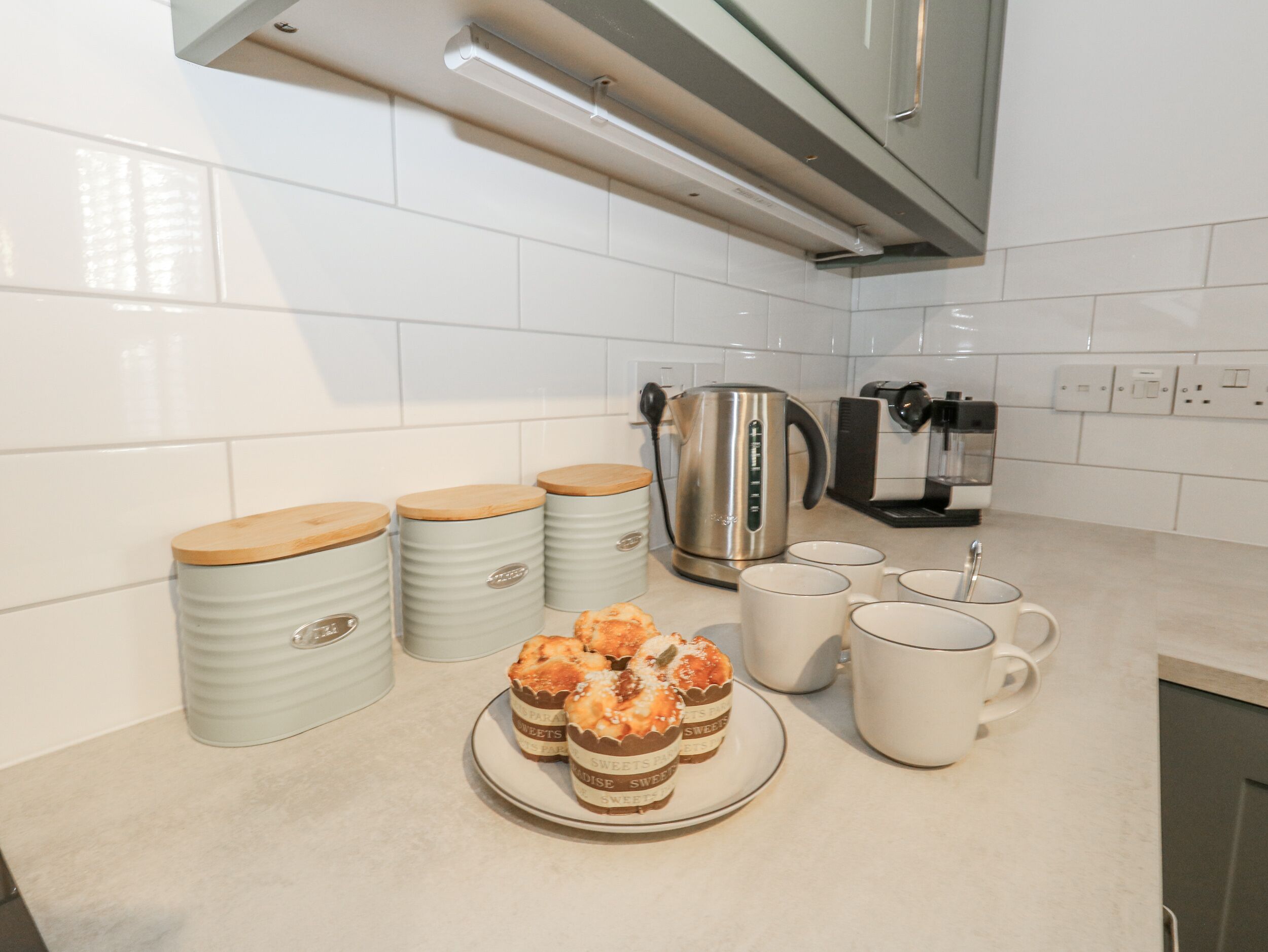 Een keuken met witte tegels, een roestvrijstalen waterkoker, een koffieapparaat en drie potten met houten deksels.