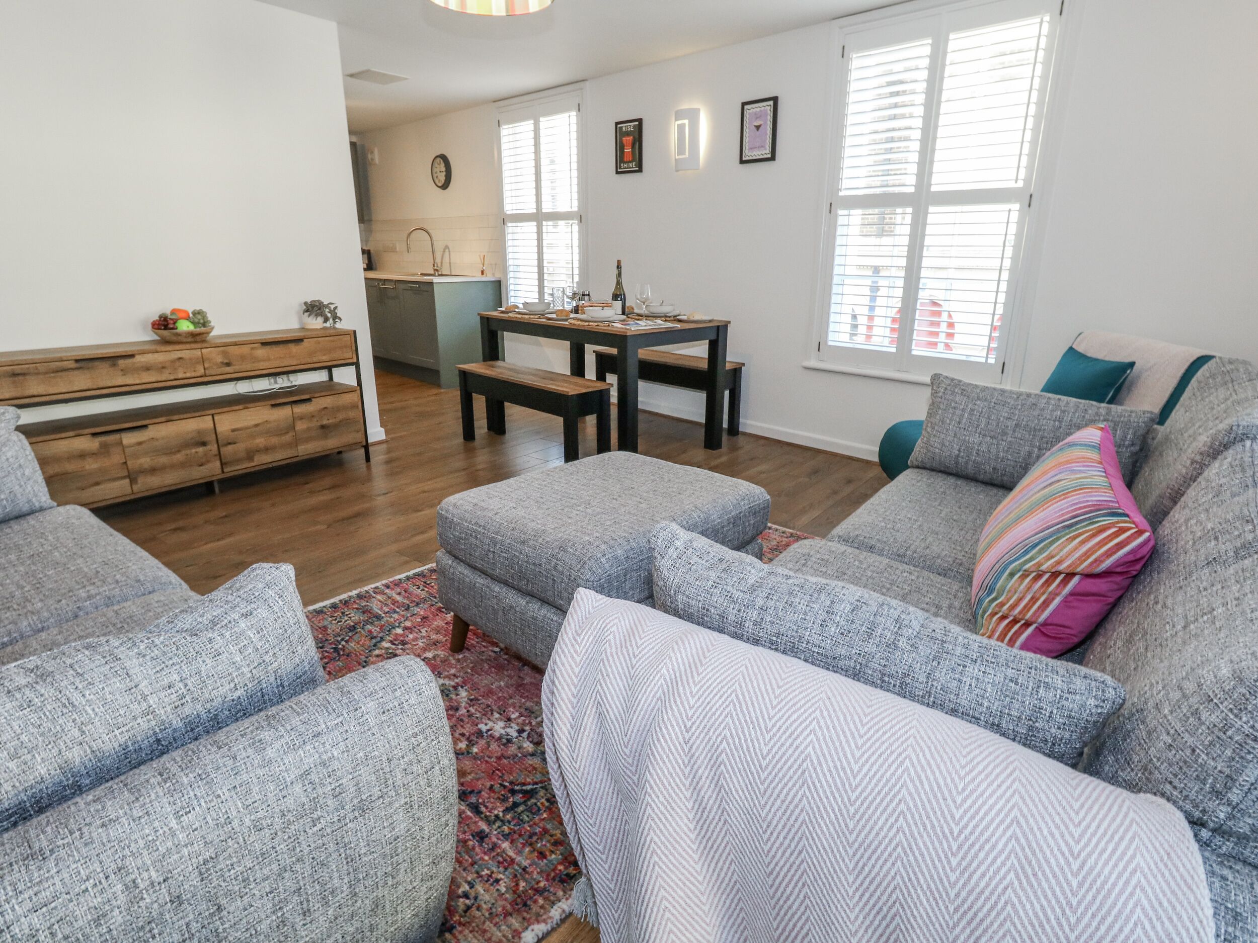 A modern living room with grey upholstered sofas, a wooden dining table, and a kitchen in the background.