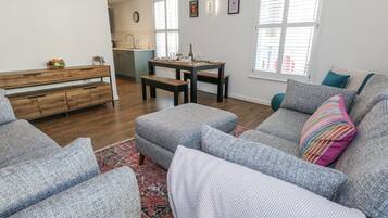 A modern living room with grey upholstered sofas, a wooden dining table, and a kitchen in the background.