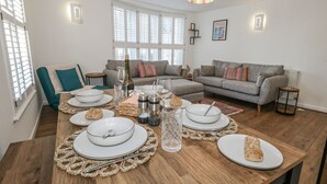 A dining area with a wooden table set for a meal, a grey sofa, and a window with blinds. - Betty's Buoy (Weymouth)