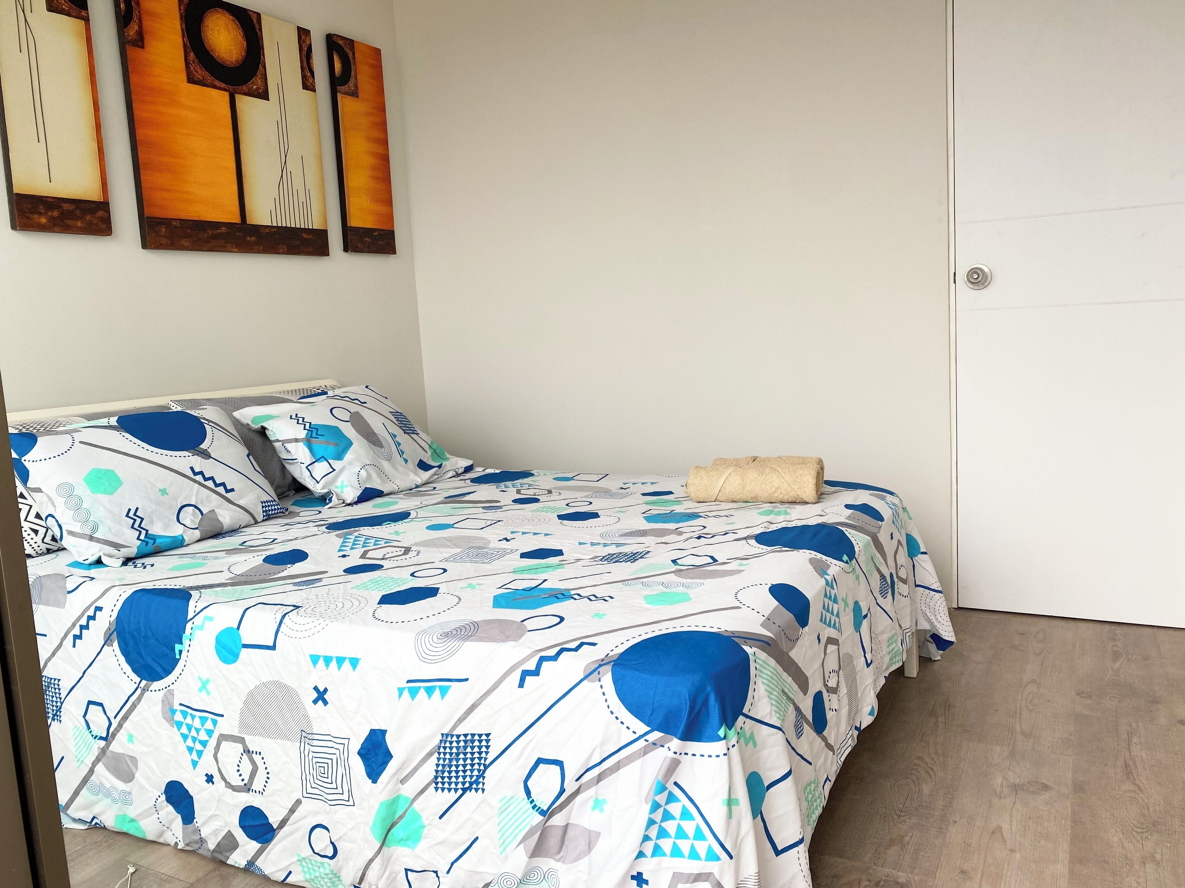 A neatly made bed with a patterned bedspread, a rolled towel, and pillows on a white wall.