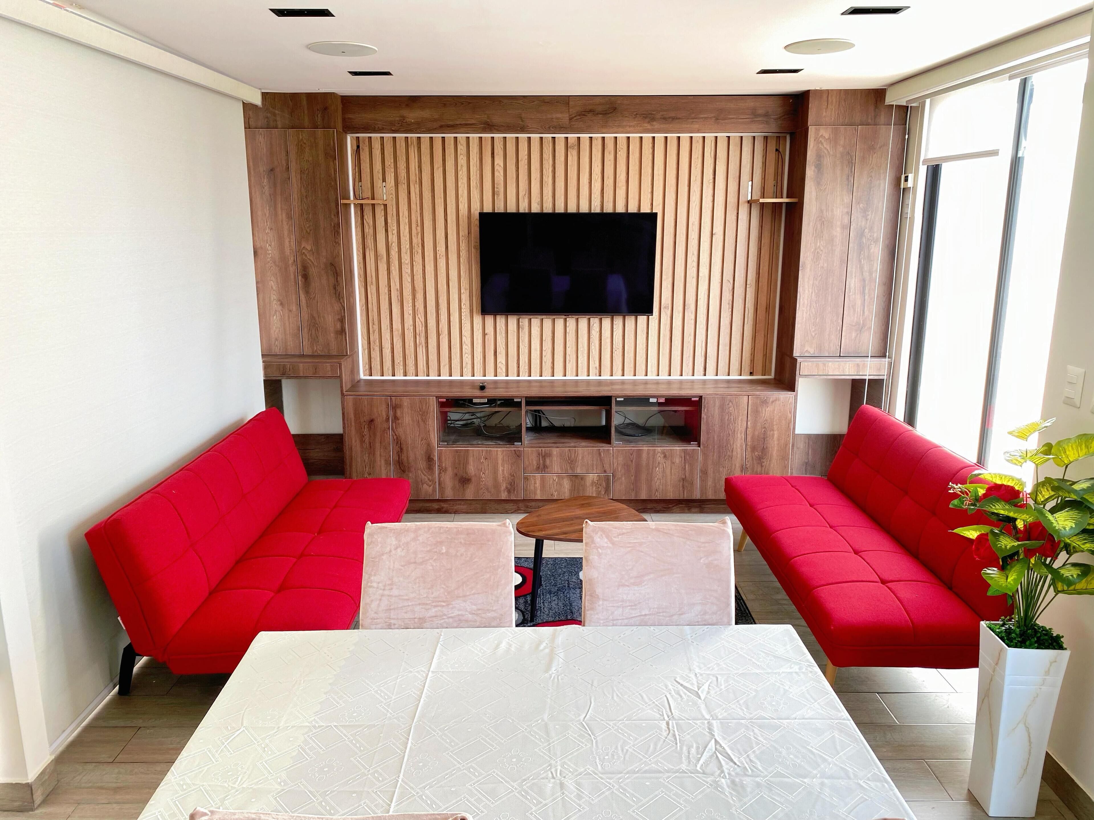 A modern living room with a flat-screen TV mounted on a wooden panel wall, two red sofas, a white table, and a potted plant.