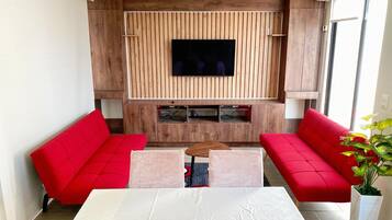 A modern living room with a flat-screen TV mounted on a wooden panel wall, two red sofas, a white table, and a potted plant.