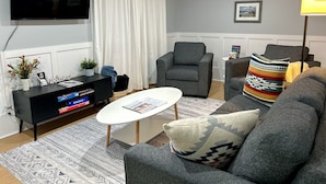 Un saló amb una televisió de pantalla plana, un sofà gris, una taula de cafè blanca, un butac armchair gris i un coixí decoratiu estampat.