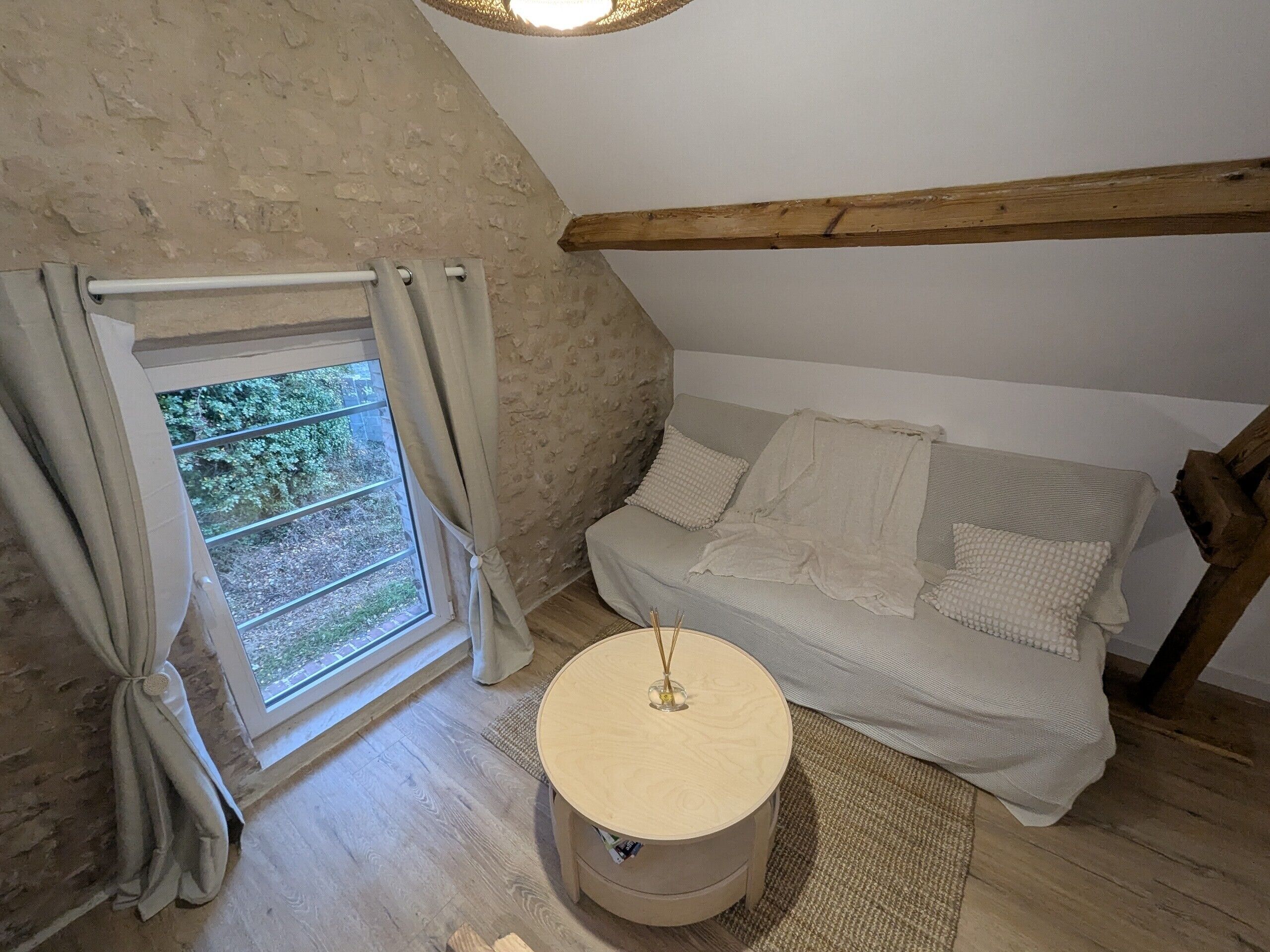 Une pièce chaleureuse avec un plafond en bois, une fenêtre avec des rideaux, un canapé avec des coussins et une petite table ronde.