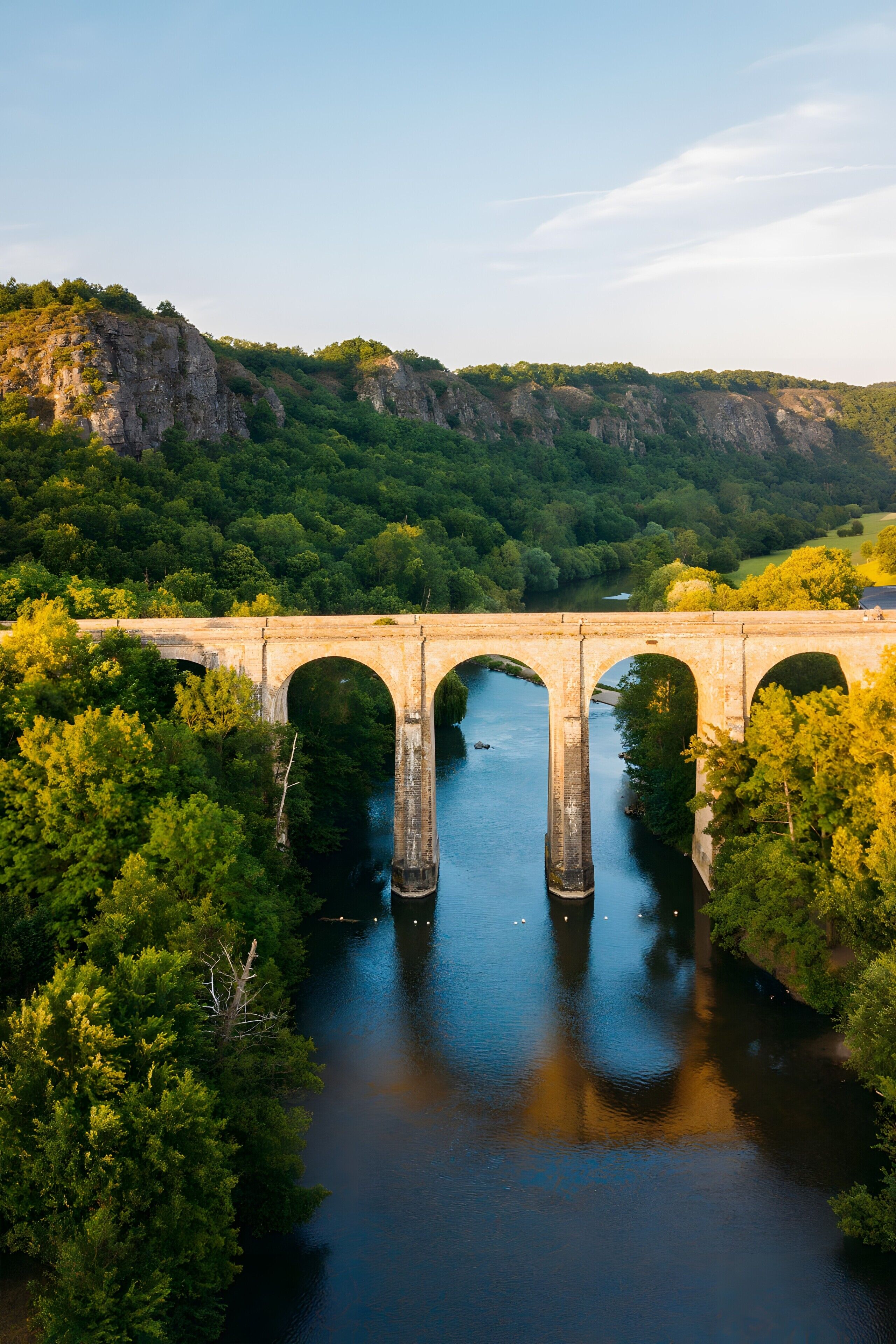 A stone bridge over a river with surrounding greenery and hills.