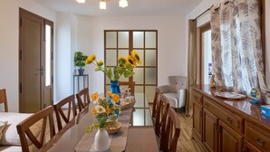 Ein Essbereich mit Tisch für vier Personen, einer Blumenvase, einer Kronleuchter und Blick durch ein mit Vorhängen drapiertes Fenster.