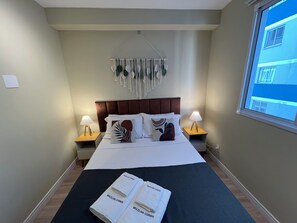 A hotel room with a bed, two bedside lamps, a macrame wall hanging, and a window with a view of buildings. - Apartamentos com piscina e academia (São Paulo)