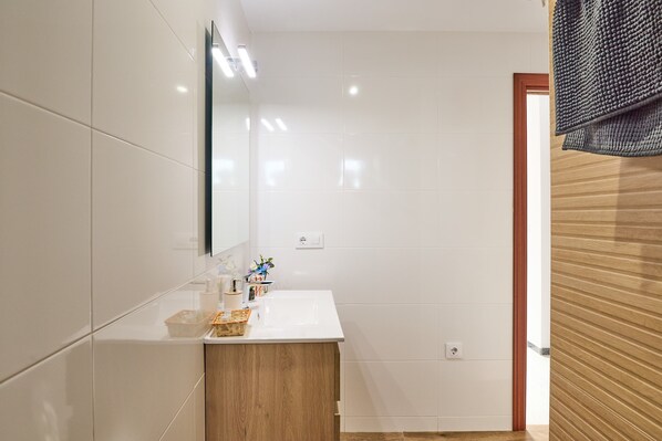 A bathroom with white tiles, a wooden vanity, a mirror, and a towel rack. - Casa Rural Emma: Mountain Views, Pool, and Air Conditioning (Zuheros)
