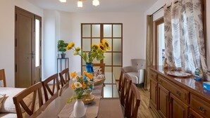 Zona pranzo con tavolo in legno, sedie, un vaso con fiori, un lampadario, una finestra con tende e un armadietto.