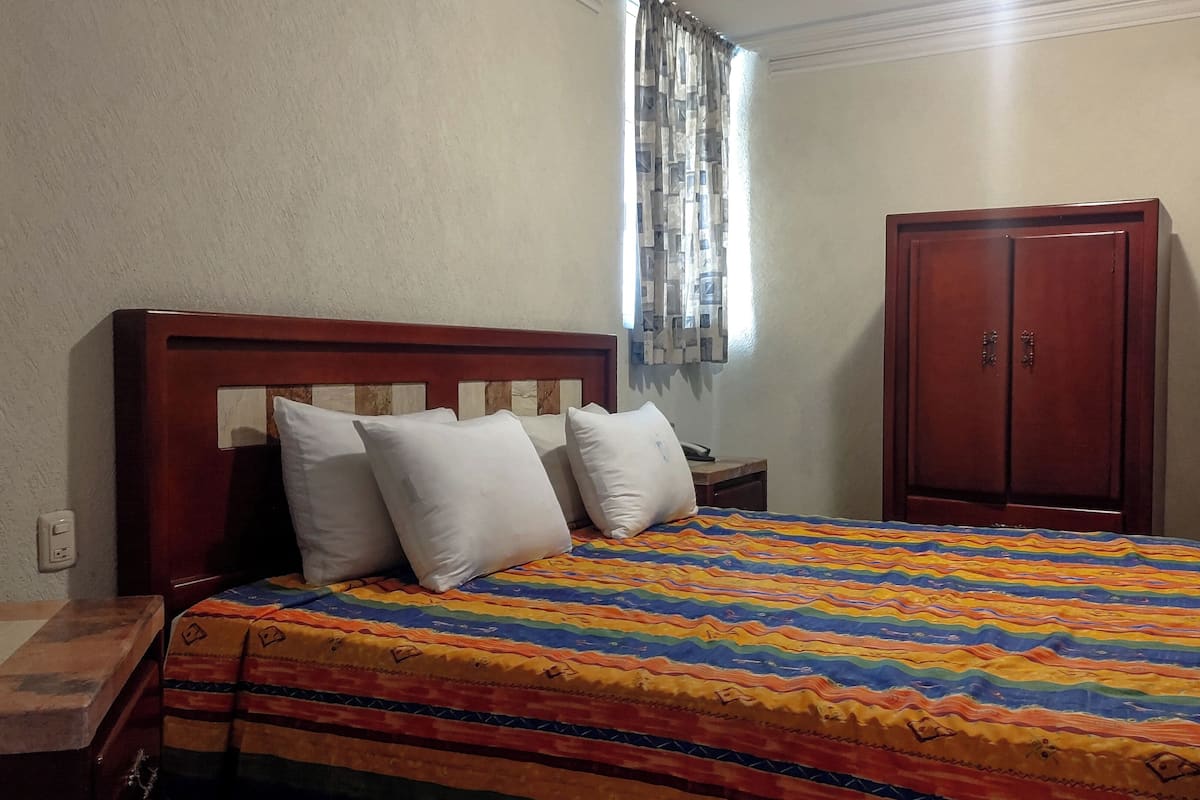 Een slaapkamer met een houten bed, witte kussens, een kleurrijk sprei, een nachtkastje van hout, een kledingkast van hout, en een raam met gordijnen.