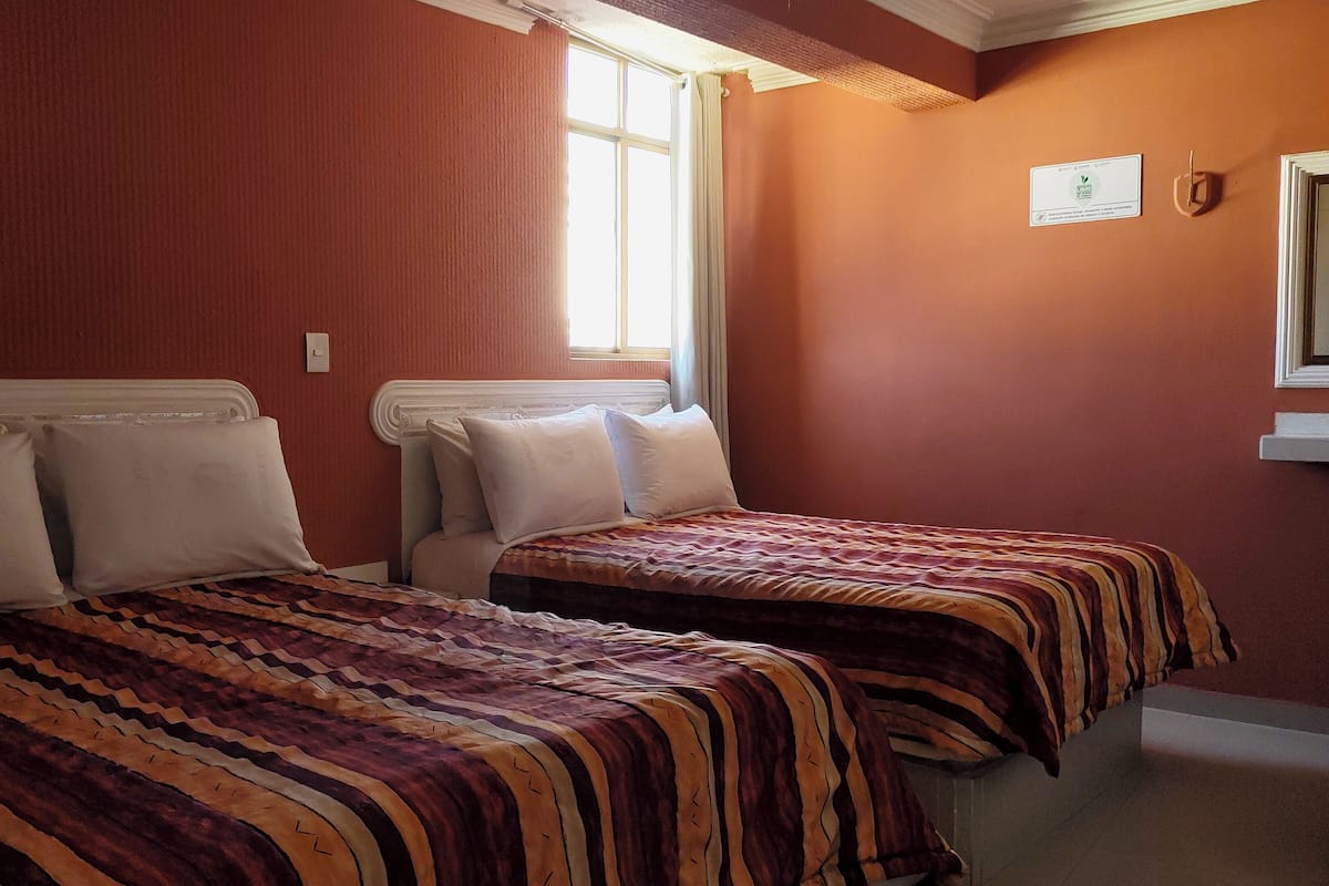 Een kamer met twee bedden, elk met een gestreepte spreidekker, een raam met gordijnen en een lichtschakelaar aan de muur.