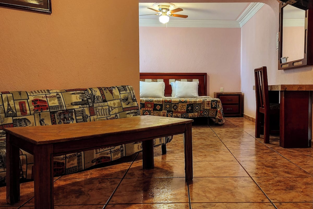 Een hotelkamer met een houten bank, een bed met beddengoed met een patroon, een kleine tafel en een plafondventilator.