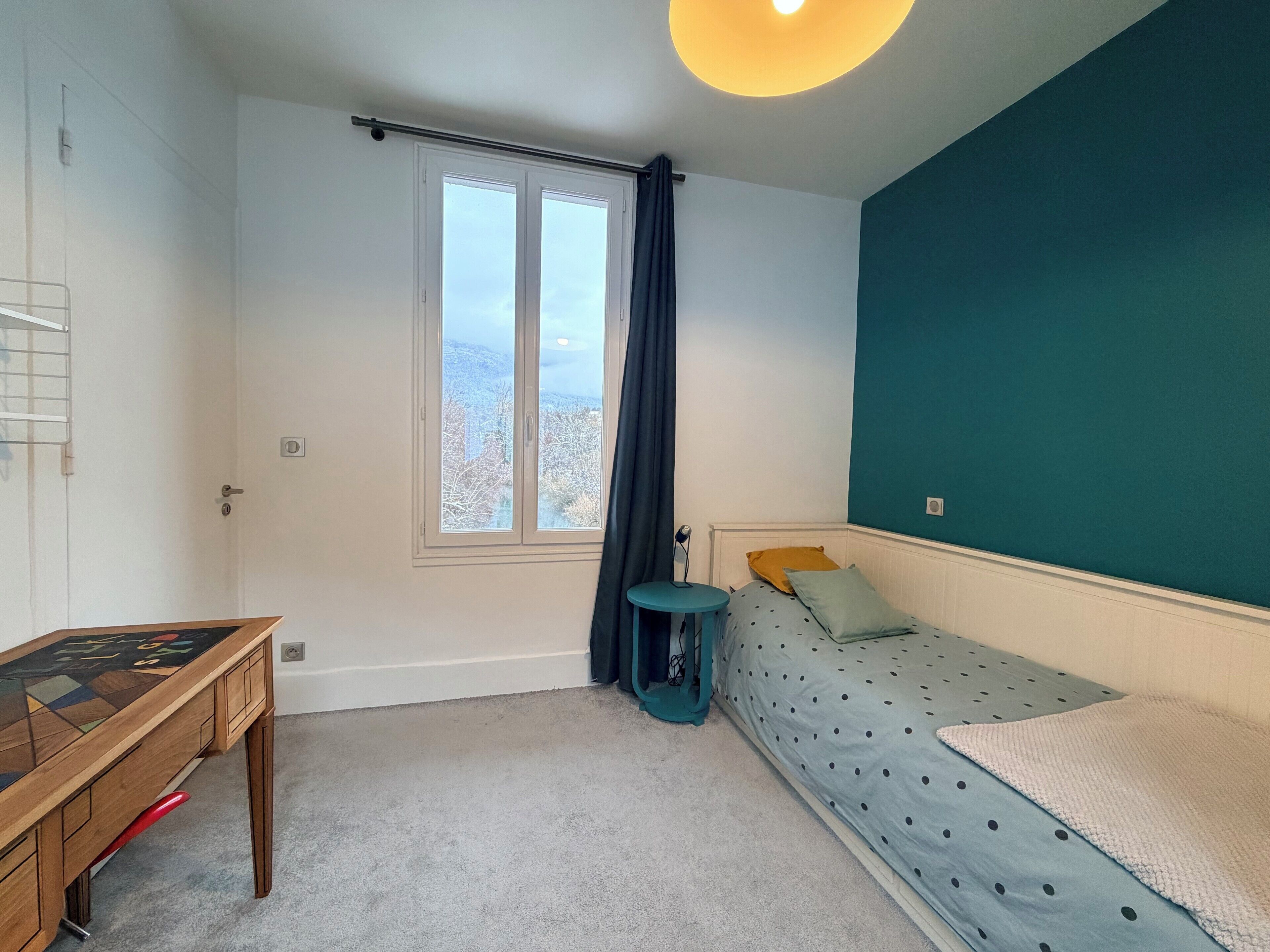 Ein Schlafzimmer mit Bett, Schreibtisch, einem Fenster mit Vorhängen und Blick auf Bäume.