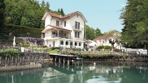 A large, two-story building with a balcony, situated next to a calm body of water, surrounded by greenery.