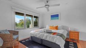 Ein Schlafzimmer mit einem Bett, einem Nachttisch, einem Korbstuhl, einem Fenster mit Blick auf Bäume und einem Deckenventilator.