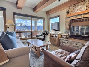 A living room with a fireplace, large windows, and a view of mountains. - The Plaza Crested Butte (Crested Butte)
