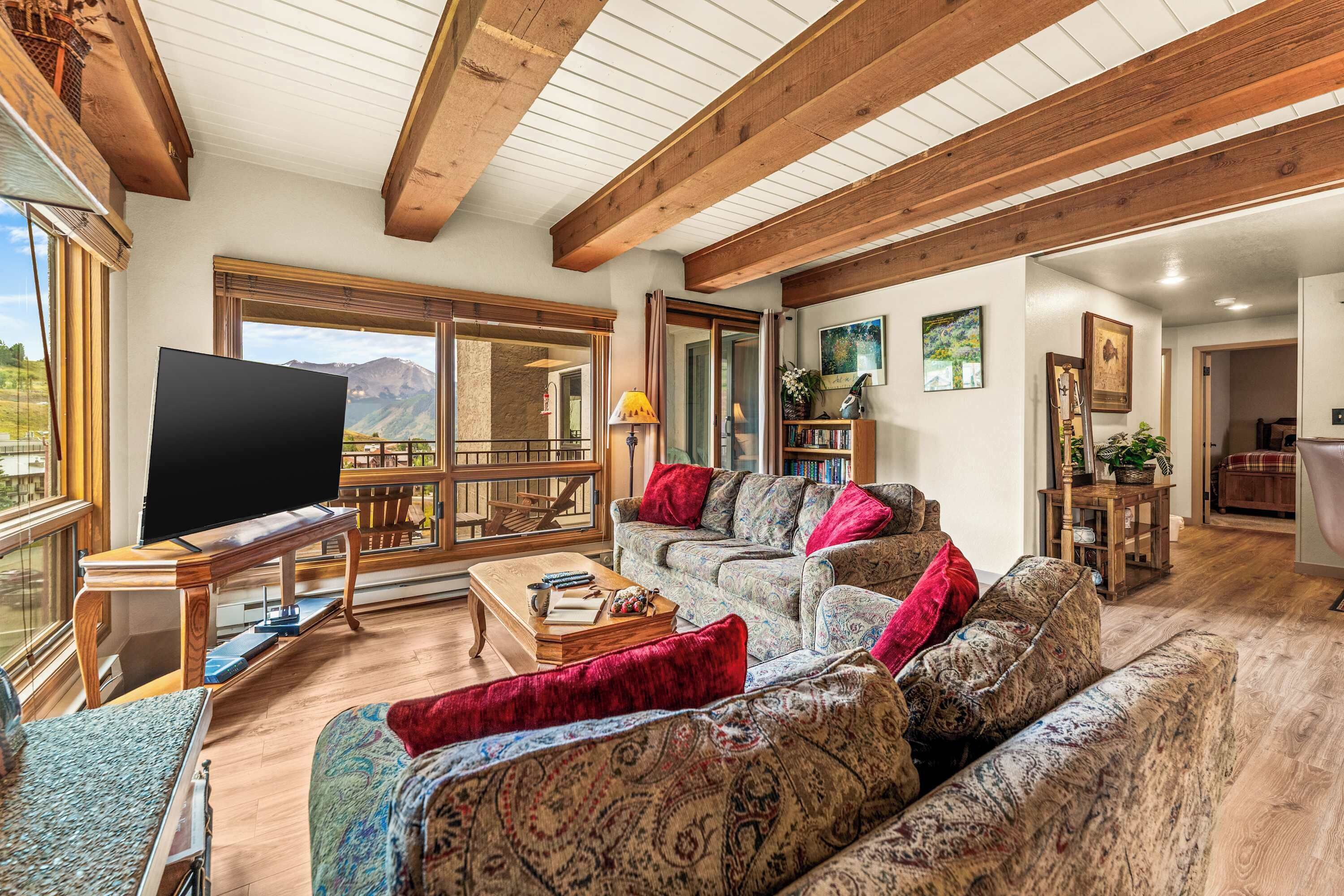 A spacious living room with a large TV, patterned sofas, wooden beams, and a view of the mountains.