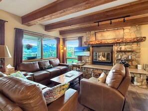 A cozy living room with a stone fireplace, leather sofas, and large windows offering scenic views. - The Plaza by Traverse Hospitality (Crested Butte)