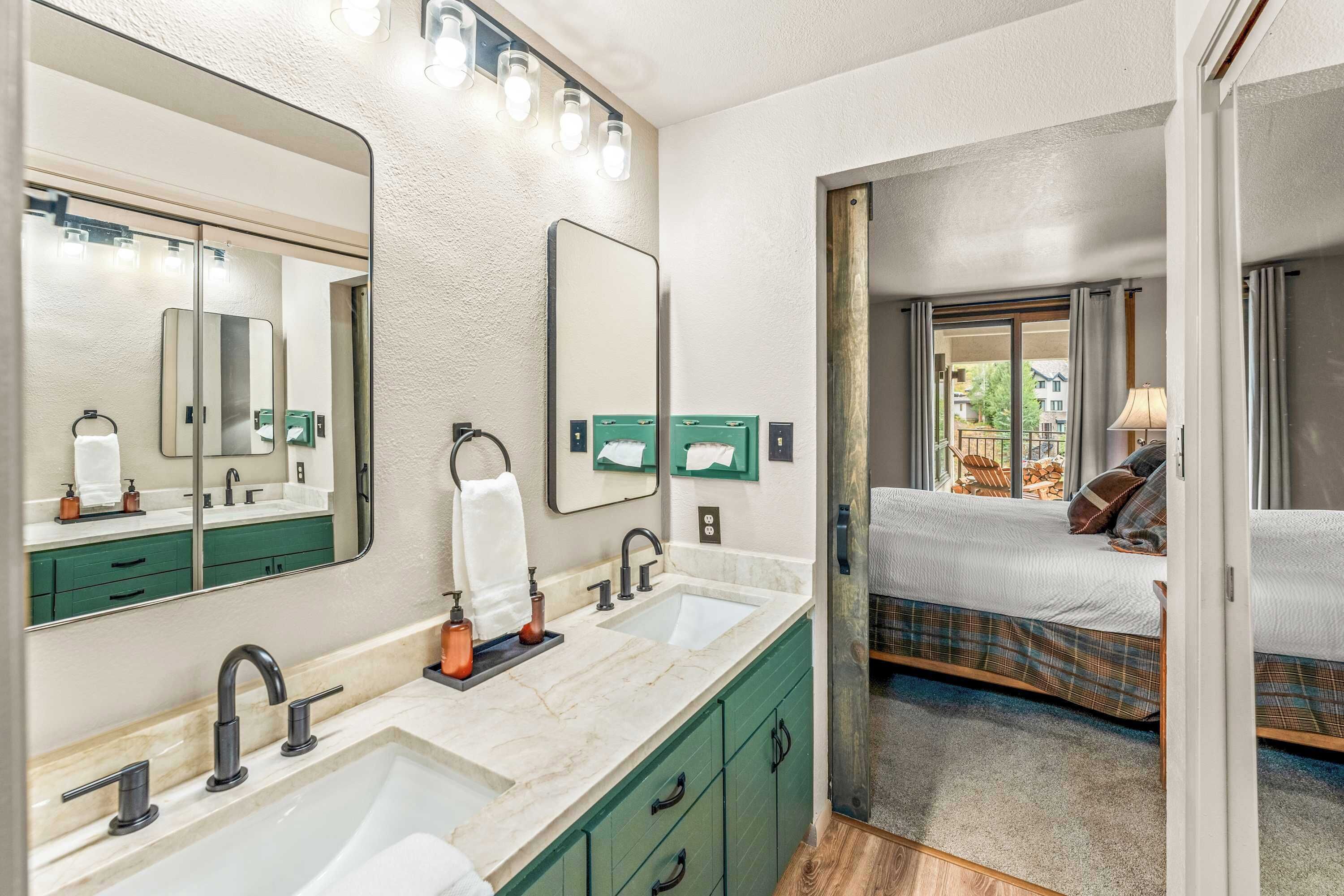Une salle de bain avec un grand miroir, deux lavabos et un lit visible à travers une porte ouverte.