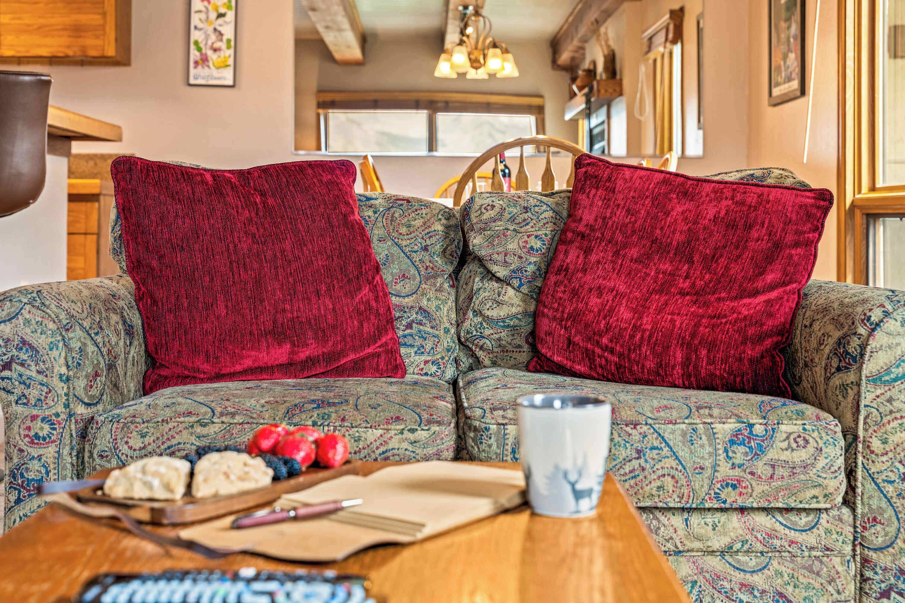 A cozy living room with a patterned sofa, red throw pillows, and a wooden coffee table with snacks and a mug.